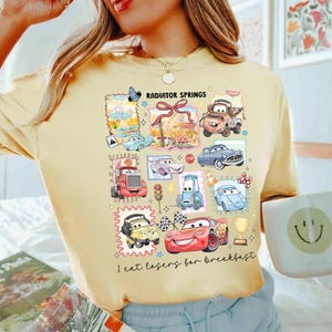 May include: Yellow t-shirt featuring a colorful graphic of various Cars characters and the text "RADIATOR SPRINGS" and "I eat losers for breakfast". The design includes cartoon cars, a trophy, and checkered flags.