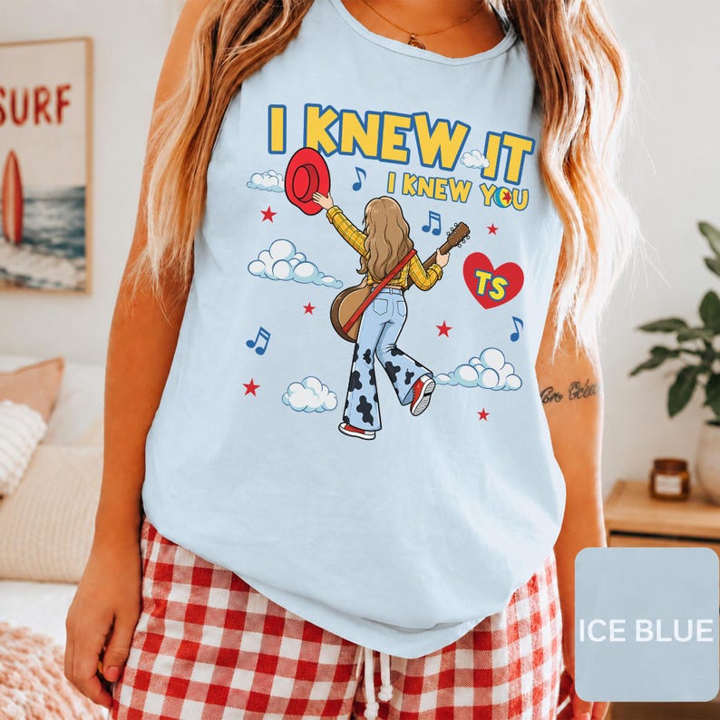 May include: A light blue tank top featuring a graphic of a person with long hair holding a red hat and a guitar. The text reads I KNEW IT I KNEW YOU with a red heart containing the letters TS, surrounded by clouds, stars, and musical notes.