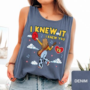 May include: A denim blue sleeveless tank top featuring a graphic illustration of a person with long hair holding a red hat and a guitar. The text reads I KNEW IT I KNEW YOU with a red heart containing the letters TS, surrounded by clouds and stars.