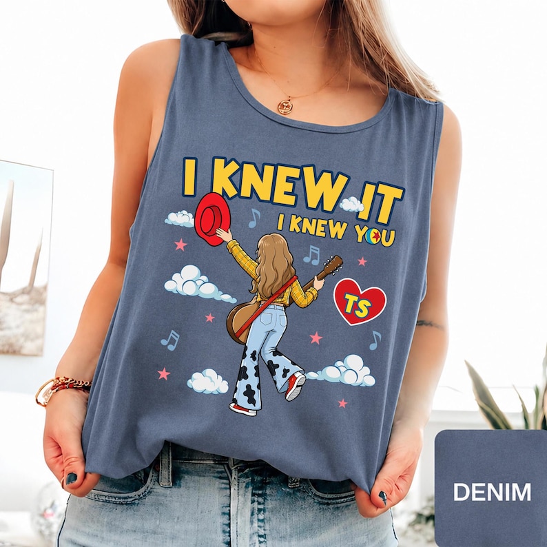 May include: A denim blue sleeveless tank top featuring a graphic illustration of a person with long hair holding a red hat and a guitar. The text reads I KNEW IT I KNEW YOU with a red heart containing the letters TS, surrounded by clouds and stars.