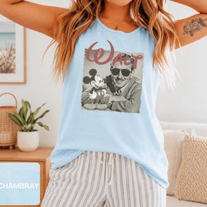 Peut inclure: Un d&eacute;bardeur bleu clair sans manches avec une impression graphique de Walt Disney portant des lunettes de soleil &agrave; c&ocirc;t&eacute; d'un Mickey Mouse en dessin anim&eacute;. Le texte "WALT" est imprim&eacute; dans une grande police stylis&eacute;e au-dessus de l'image.