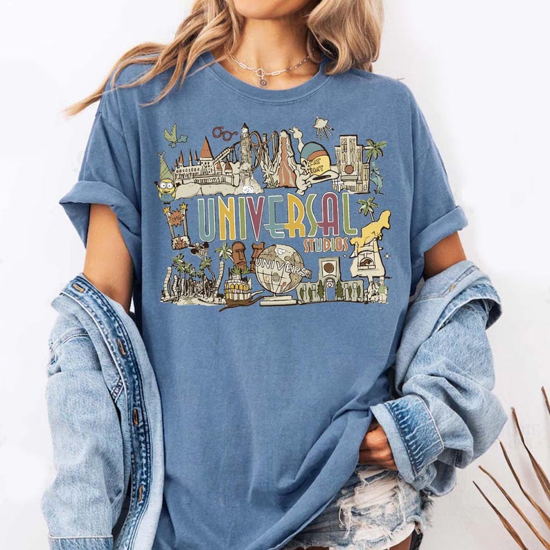 May include: A blue t-shirt with a colourful graphic of Universal Studios attractions and the text "UNIVERSAL STUDIOS". The design includes various landmarks and characters, creating a vintage, travel-themed aesthetic.