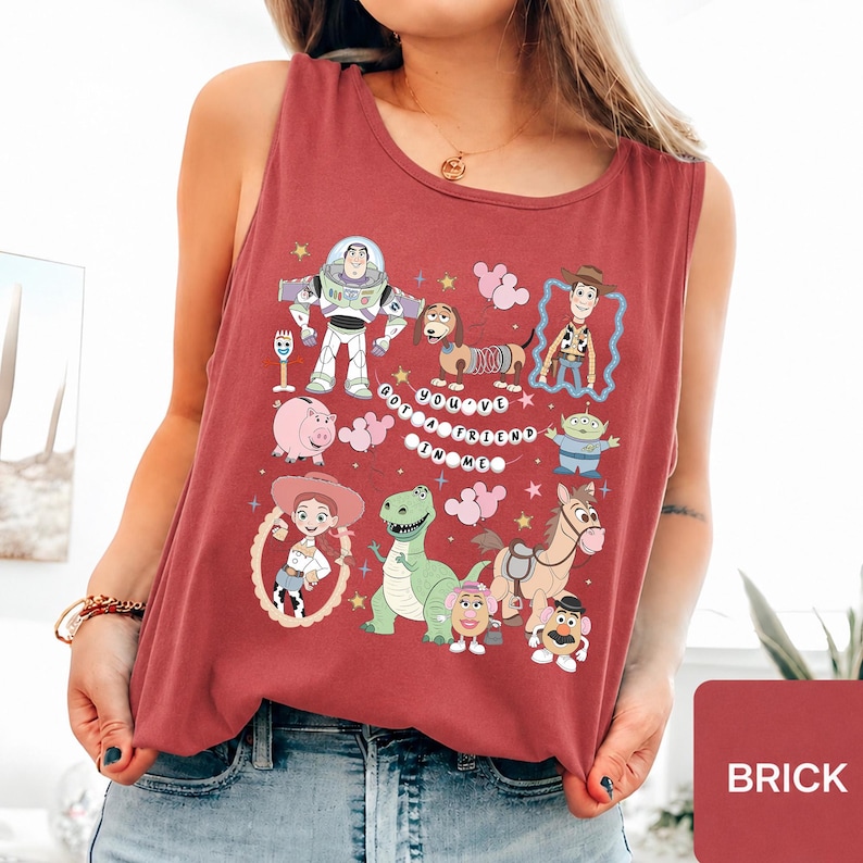 May include: A brick red sleeveless tank top featuring a graphic print of various Toy Story characters including Buzz Lightyear, Woody, Jessie, Rex, and Slinky Dog, with the text "YOU'VE GOT A FRIEND IN ME" arranged in a circular pattern.