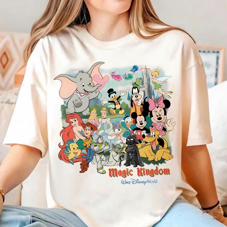 K&ouml;nnte beinhalten: Cremefarbenes T-Shirt mit einem farbenfrohen Aufdruck verschiedener Disney-Figuren, darunter Dumbo, Mickey Mouse und Arielle. Die Worte "Magic Kingdom" und "Walt Disney World" sind darunter gedruckt. Das T-Shirt hat kurze &Auml;rmel.