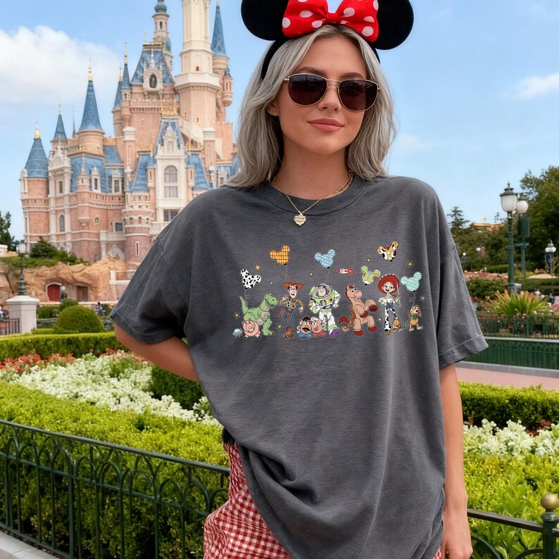 K&ouml;nnte beinhalten: Ein anthrazitfarbenes Kurzarm-T-Shirt mit einem bunten Grafikdruck verschiedener animierter Spielzeugfiguren, die in einer Reihe stehen und jeweils einen gemusterten Luftballon halten. Zu sehen sind ein gr&uuml;ner Dinosaurier, ein Cowboy und eine Cowgirl.