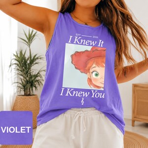 May include: A violet sleeveless tank top featuring a graphic print of a cartoon character with red hair and a red hat. The text reads EST 2026 I Knew It I Knew You with a small musical note symbol below the text. The shirt has a round neckline.
