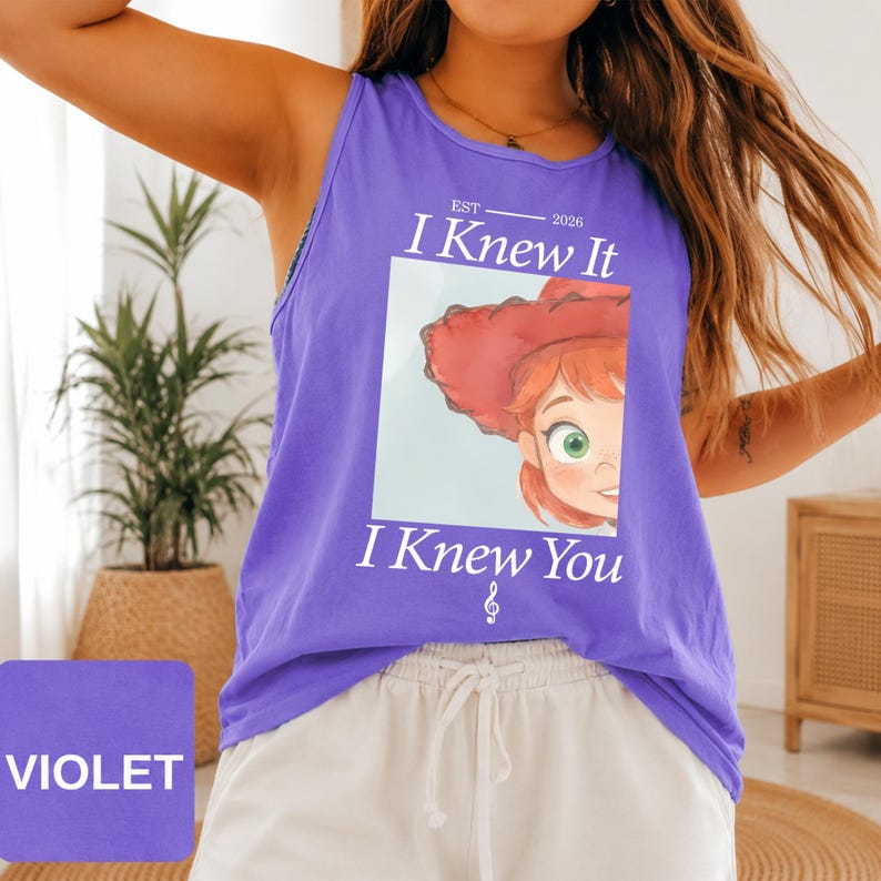 May include: A violet sleeveless tank top featuring a graphic print of a cartoon character with red hair and a red hat. The text reads EST 2026 I Knew It I Knew You with a small musical note symbol below the text. The shirt has a round neckline.