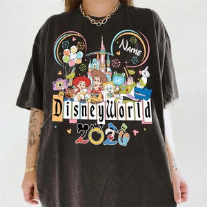 Peut inclure: T-shirt gris fonc&eacute; avec un motif color&eacute; de Disney World. Il pr&eacute;sente des personnages de dessins anim&eacute;s, un ch&acirc;teau et le texte "Disneyworld 2020". Comprend des oreilles de Mickey et un espace pour un nom.