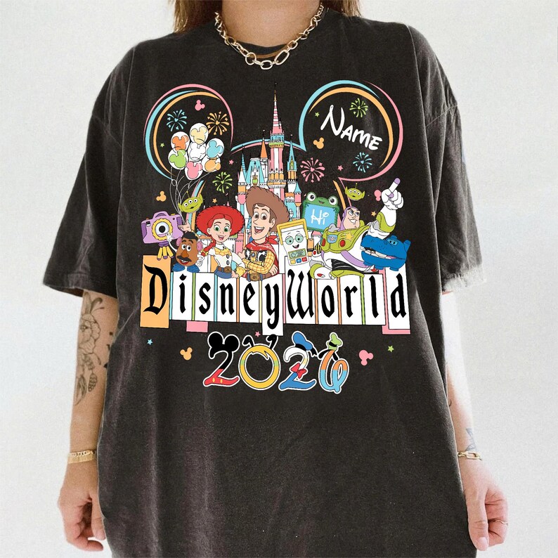 Peut inclure: T-shirt gris fonc&eacute; avec un motif color&eacute; de Disney World. Il pr&eacute;sente des personnages de dessins anim&eacute;s, un ch&acirc;teau et le texte "Disneyworld 2020". Comprend des oreilles de Mickey et un espace pour un nom.