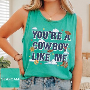May include: A seafoam green sleeveless tank top featuring the dark blue text "YOU'RE A COWBOY LIKE ME" surrounded by graphic illustrations of a brown cowboy hat, a sheriff badge, a red bandana, a ball, and brown cowboy boots with white clouds.