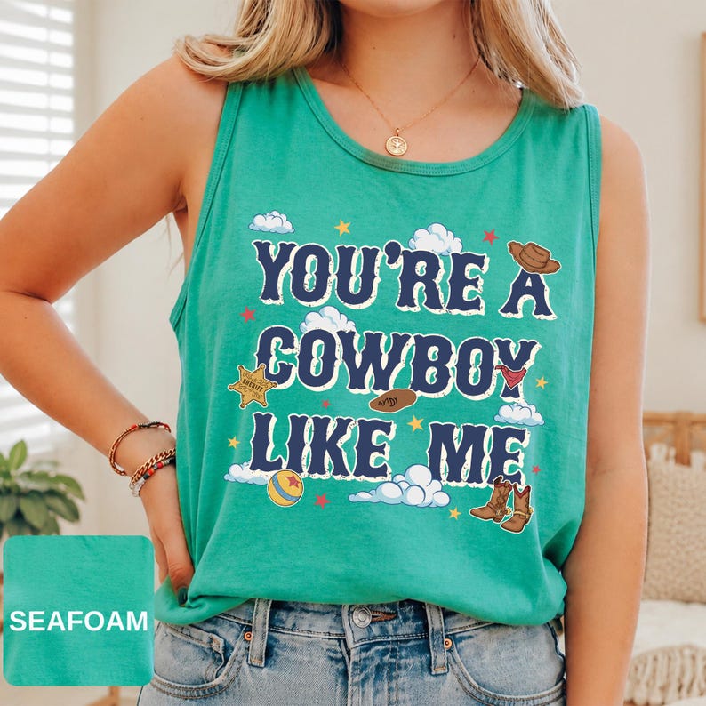 May include: A seafoam green sleeveless tank top featuring the dark blue text "YOU'RE A COWBOY LIKE ME" surrounded by graphic illustrations of a brown cowboy hat, a sheriff badge, a red bandana, a ball, and brown cowboy boots with white clouds.