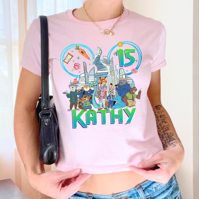 May include: A light pink t-shirt featuring a colorful cartoon graphic with the text "KATHY" in green. The graphic includes a cityscape, the number "15", and various animal characters. The shirt has short sleeves and a crew neck.