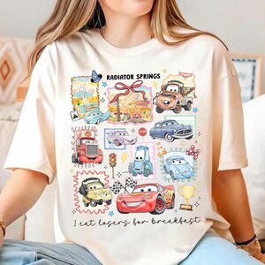 May include: Cream-colored t-shirt featuring a colorful graphic of various cartoon cars and the text "Radiator Springs" and "I eat lasers for breakfast". The design includes a variety of car characters and related imagery.