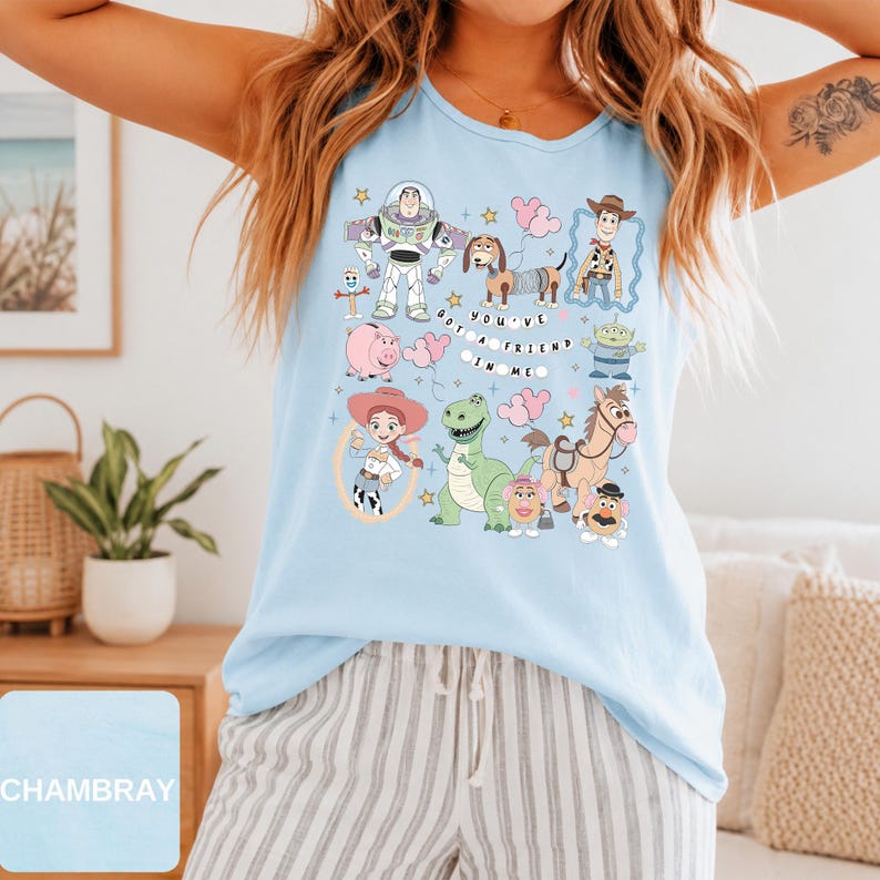 May include: A light blue sleeveless tank top featuring a graphic print of various animated characters including a space ranger, a cowboy, a cowgirl, a dinosaur, and a dog. The text "YOU'VE GOT A FRIEND IN ME" is printed in the center.