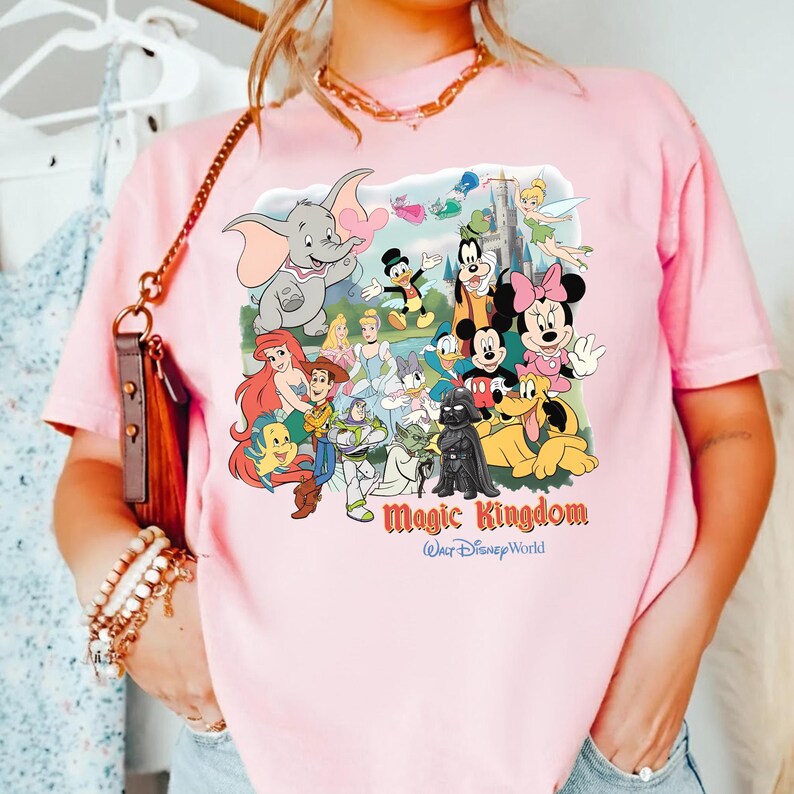 K&ouml;nnte beinhalten: Hellrosa T-Shirt mit einem farbenfrohen Aufdruck von Disney-Figuren, darunter Mickey Mouse, Minnie Mouse und Dumbo. Der Aufdruck enth&auml;lt den Text "Magic Kingdom" und "Walt Disney World".