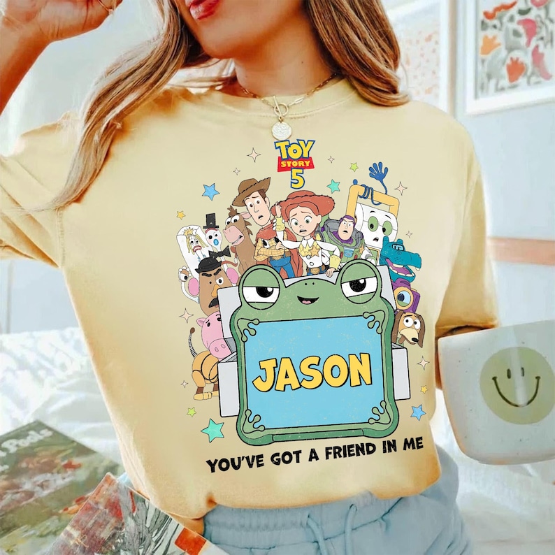 May include: Yellow t-shirt featuring a colorful graphic of Toy Story characters, the text "TOY STORY 5", and a frog holding a sign that says "JASON". The phrase "YOU'VE GOT A FRIEND IN ME" is printed at the bottom.