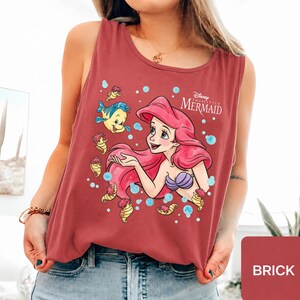 Peut inclure: Un d&eacute;bardeur sans manches rouge brique avec une impression graphique d'Ariel de La Petite Sir&egrave;ne, ses longs cheveux rouges, un haut coquillage violet et le poisson Polochon, entour&eacute;s de petits hippocampes jaunes et de bulles bleu clair.