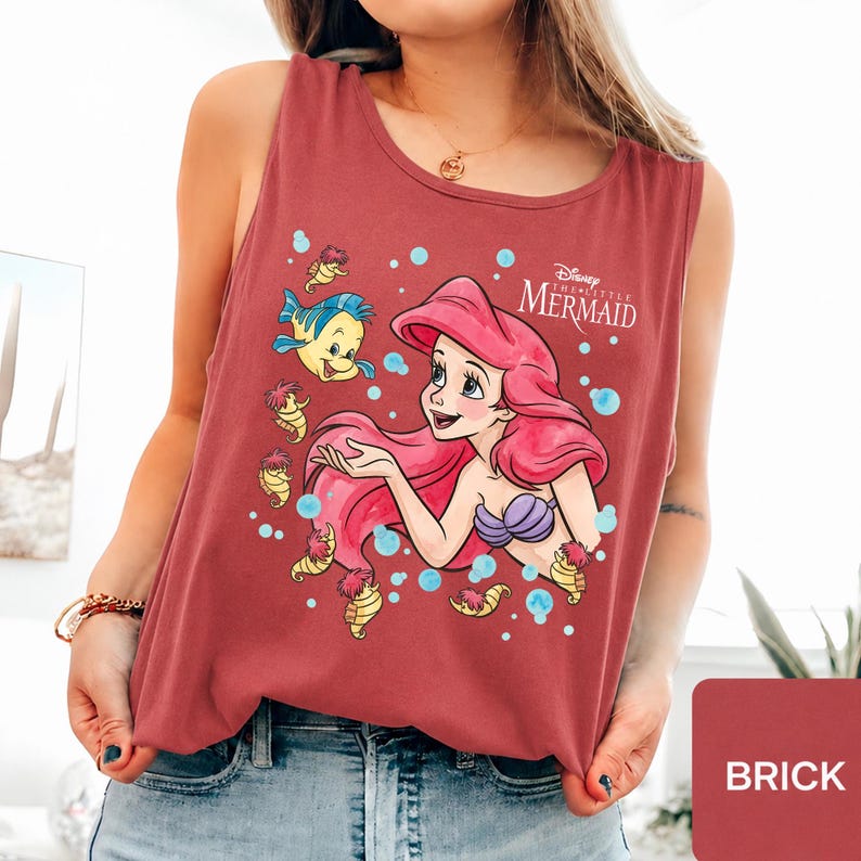 Peut inclure: Un d&eacute;bardeur sans manches rouge brique avec une impression graphique d'Ariel de La Petite Sir&egrave;ne, ses longs cheveux rouges, un haut coquillage violet et le poisson Polochon, entour&eacute;s de petits hippocampes jaunes et de bulles bleu clair.
