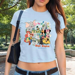 K&ouml;nnte beinhalten: Hellblaues T-Shirt mit einer farbenfrohen Grafik von Disney-Figuren, darunter Mickey und Minnie Mouse, Dumbo und andere. Die Worte "Magic Kingdom" sind unter den Figuren aufgedruckt. Das Shirt hat kurze &Auml;rmel.