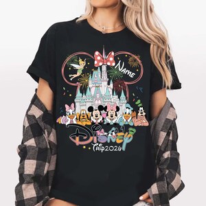 May include: Black t-shirt featuring a colorful Disney design. The design includes the Disney castle, various Disney characters, fireworks, and the text "Disney Trip 2026". A pink bow and the word "Name" are also included.