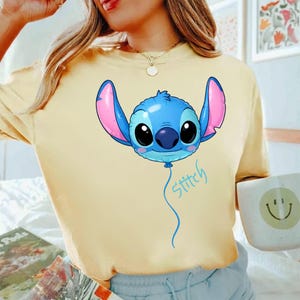 May include: A pale yellow t-shirt featuring a graphic of the character Stitch from Lilo and Stitch depicted as a blue balloon with large pink ears and black eyes. The word Stitch is written in a light blue cursive script below the balloon.