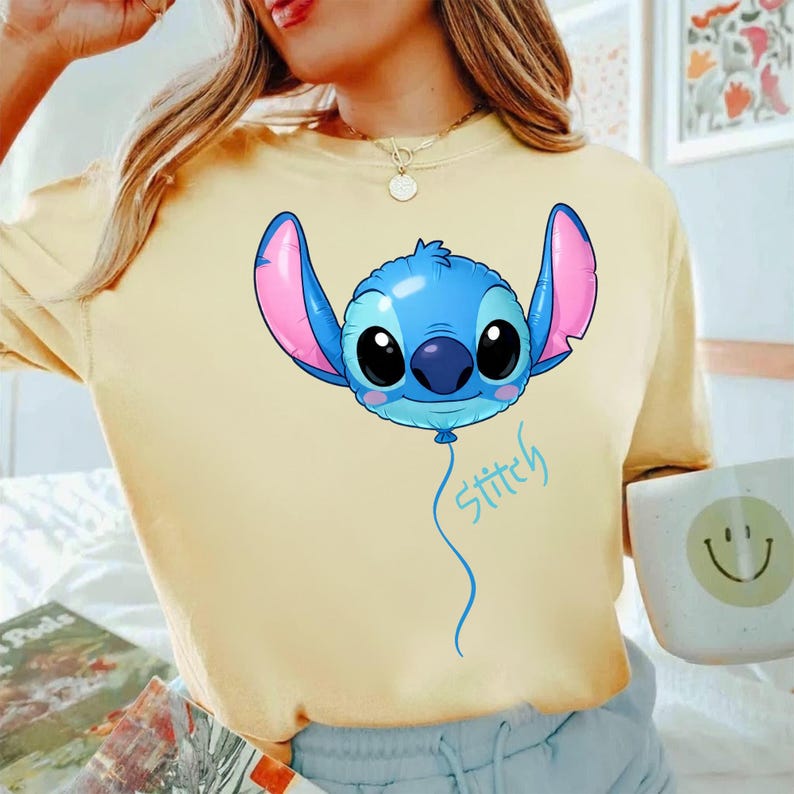 May include: A pale yellow t-shirt featuring a graphic of the character Stitch from Lilo and Stitch depicted as a blue balloon with large pink ears and black eyes. The word Stitch is written in a light blue cursive script below the balloon.
