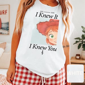 May include: A white sleeveless tank top featuring a graphic print of a cartoon character with red hair and a red hat. The text reads "EST 2026 I Knew It I Knew You" with a small musical note symbol below the text, centered on the front of the garment.