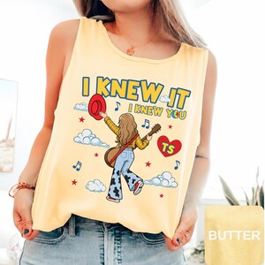 May include: A pale yellow sleeveless tank top featuring a graphic illustration of a person with long hair holding a red hat and a guitar. The text reads I KNEW IT I KNEW YOU with blue musical notes, white clouds, red stars, and a red heart with TS.