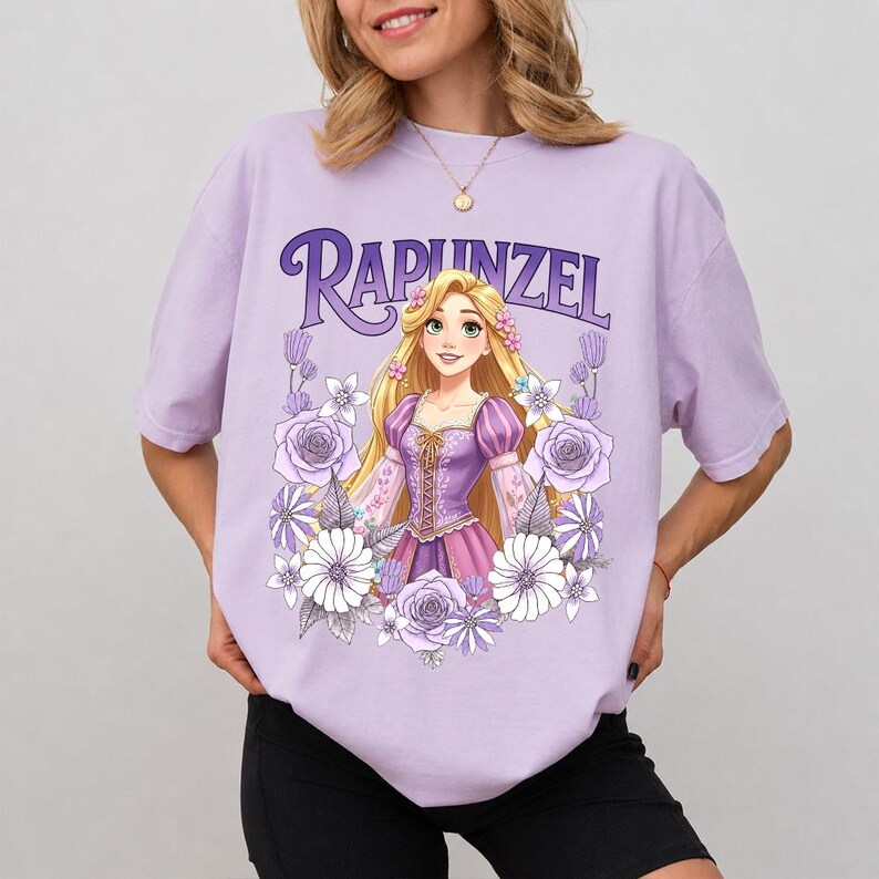 Puede incluir: Una camiseta lila claro con la palabra "Rapunzel" en una fuente estilizada sobre una ilustraci&oacute;n de dibujos animados de una princesa rubia con un vestido morado, rodeada de flores. La camiseta es de material suave.
