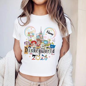 Peut inclure: T-shirt blanc avec un motif color&eacute; de Disney World. Le design comprend le texte "Disneyworld" dans une police ludique, l'ann&eacute;e "2020" et divers personnages Disney. Un nom personnalisable est &eacute;galement inclus.