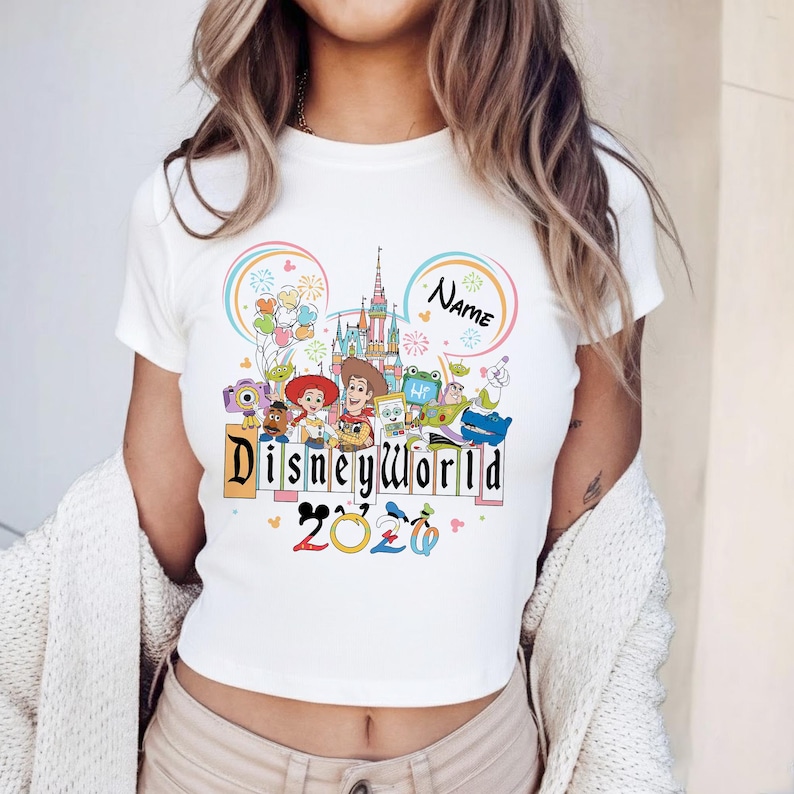 Peut inclure: T-shirt blanc avec un motif color&eacute; de Disney World. Le design comprend le texte "Disneyworld" dans une police ludique, l'ann&eacute;e "2020" et divers personnages Disney. Un nom personnalisable est &eacute;galement inclus.
