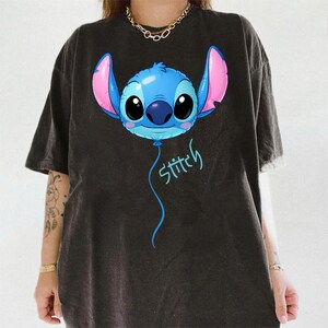 May include: A black short-sleeved t-shirt featuring a graphic of a blue character head shaped like a balloon with large black eyes, pink inner ears, and a blue nose. Below the character, the word Stitch is written in a light blue script font.