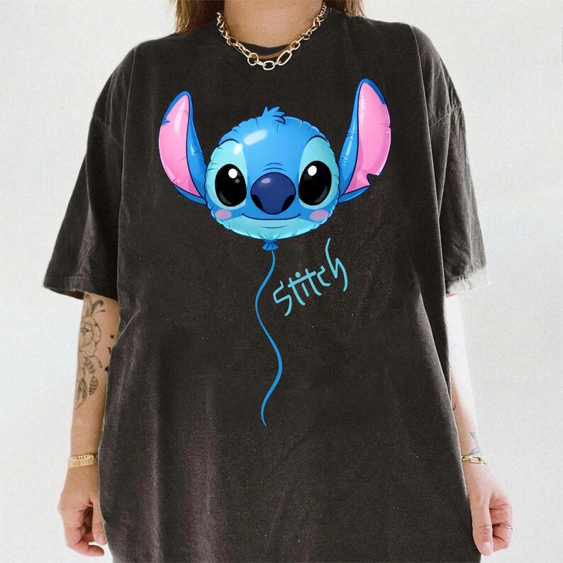 May include: A black short-sleeved t-shirt featuring a graphic of a blue character head shaped like a balloon with large black eyes, pink inner ears, and a blue nose. Below the character, the word Stitch is written in a light blue script font.