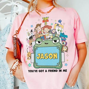 May include: Pink t-shirt featuring a colorful graphic of Toy Story characters. The design includes the text "TOY STORY 5" and a frog holding a sign that says "JASON". The phrase "YOU'VE GOT A FRIEND IN ME" is also printed on the shirt.