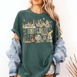 May include: Green t-shirt featuring a graphic of Universal Studios attractions. The design includes the text "UNIVERSAL STUDIOS" in a colourful, vintage style, with various theme park icons. The shirt has short sleeves and a relaxed fit.