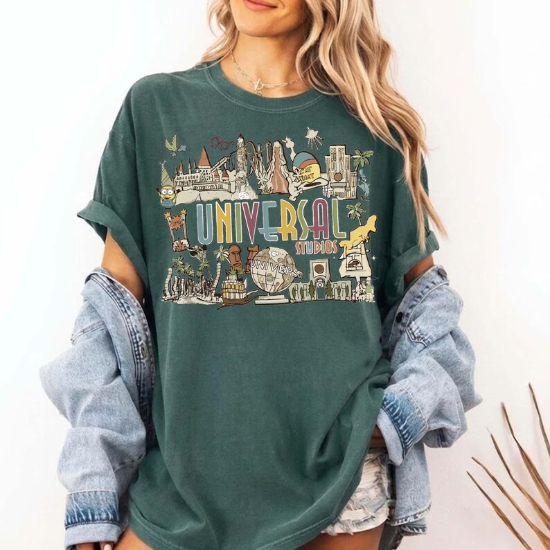 May include: Green t-shirt featuring a graphic of Universal Studios attractions. The design includes the text "UNIVERSAL STUDIOS" in a colourful, vintage style, with various theme park icons. The shirt has short sleeves and a relaxed fit.