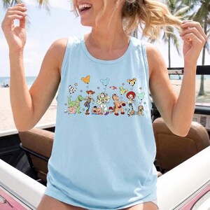 May include: Light blue tank top featuring a colorful graphic of cartoon characters and balloons. The design includes a dinosaur, cowboy, space ranger, horse, and other characters. The tank top is made of a soft material.