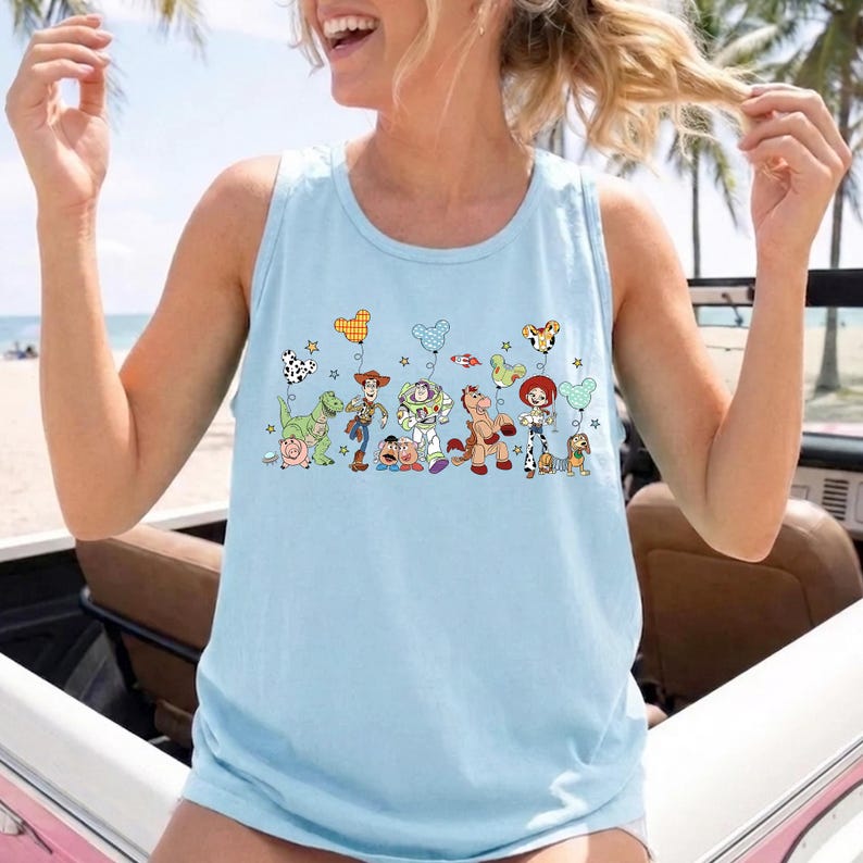 May include: Light blue tank top featuring a colorful graphic of cartoon characters and balloons. The design includes a dinosaur, cowboy, space ranger, horse, and other characters. The tank top is made of a soft material.