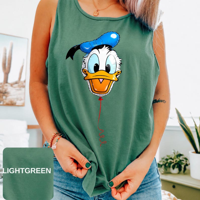 Op de afbeelding: Een lichtgroene mouwloze tanktop met een grafische print van Donald Duck ontworpen als een blauw-witte ballon. Het personage heeft een gele snavel en zit vast aan een dun rood touwtje dat naar beneden loopt op de voorkant van het kledingstuk.
