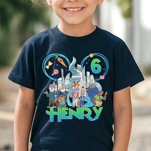 May include: Navy blue t-shirt featuring a colorful graphic of cartoon characters and the number 6. The design includes the name "HENRY" in a gradient font, along with city buildings and other cartoon elements. The shirt is designed for a child.
