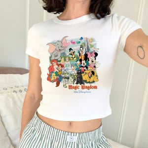 K&ouml;nnte beinhalten: Wei&szlig;es Crop-Top mit einer farbenfrohen Grafik von Disney-Figuren, darunter Mickey Mouse, Minnie Mouse und Dumbo. Der Text "Magic Kingdom" ist in einer stilisierten Schriftart gedruckt. Das Shirt hat kurze &Auml;rmel.