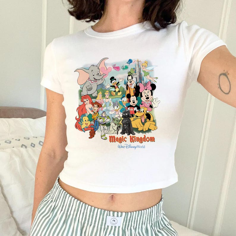 K&ouml;nnte beinhalten: Wei&szlig;es Crop-Top mit einer farbenfrohen Grafik von Disney-Figuren, darunter Mickey Mouse, Minnie Mouse und Dumbo. Der Text "Magic Kingdom" ist in einer stilisierten Schriftart gedruckt. Das Shirt hat kurze &Auml;rmel.