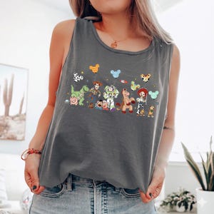 May include: A charcoal gray tank top featuring a colorful graphic of animated characters. The design includes a dinosaur, a pig, Woody, Buzz Lightyear, Jessie, and other characters. The tank top is sleeveless and made of a soft material.