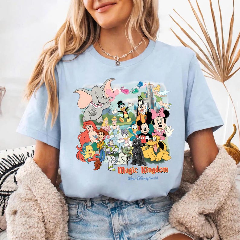 K&ouml;nnte beinhalten: Hellblaues T-Shirt mit einem farbenfrohen Aufdruck von Disney-Figuren, darunter Mickey Mouse, Minnie Mouse und Dumbo. Der Text "Magic Kingdom" ist unter den Figuren aufgedruckt. Das Shirt besteht aus einem weichen Material.