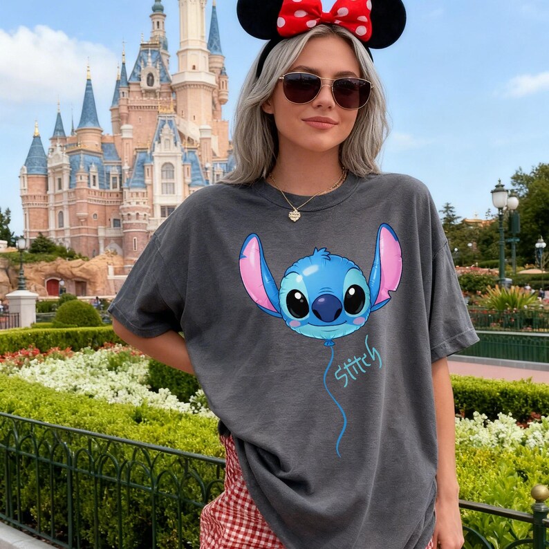 May include: A charcoal gray short-sleeve t-shirt featuring a graphic of the character Stitch designed as a blue balloon with large ears and pink inner accents. The word Stitch is written in a light blue script font below the balloon graphic.