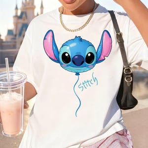 May include: A white short-sleeved t-shirt featuring a graphic of a blue Stitch character designed as a balloon with large pink-lined ears and black eyes. Below the balloon, the word Stitch is written in a light blue cursive script font.