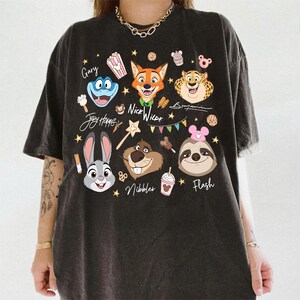 Pu&ograve; includere: T-shirt grigio scuro con personaggi dei cartoni animati di Zootropolis. Il design include Judy Hopps, Nick Wilde, Flash e altri personaggi, con i loro nomi. La maglietta ha un aspetto vintage e usurato.
