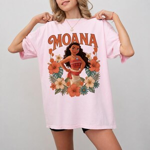 Puede incluir: Camiseta rosa claro con la palabra "MOANA" en letras naranjas sobre un dibujo de Moana rodeada de flores. El dibujo incluye flores naranjas, blancas y azules y hojas verdes. La camiseta es de corte holgado.