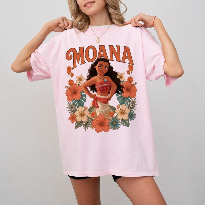 Puede incluir: Camiseta rosa claro con la palabra "MOANA" en letras naranjas sobre un dibujo de Moana rodeada de flores. El dibujo incluye flores naranjas, blancas y azules y hojas verdes. La camiseta es de corte holgado.