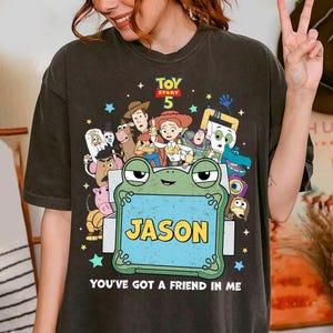 May include: Dark gray t-shirt featuring a colorful graphic of characters from Toy Story 5. The design includes a frog holding a sign with the name "JASON" and the phrase "YOU'VE GOT A FRIEND IN ME".